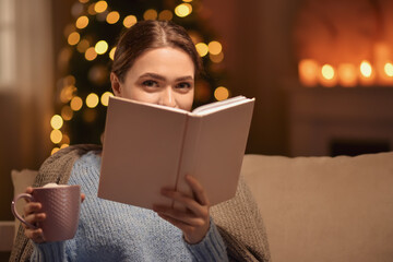 Beautiful young woman drinking hot chocolate and reading book at home on Christmas eve