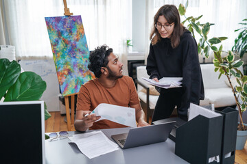 Caucasian female stylist and her indian coworker discuss about their project in modern office room in daytime.