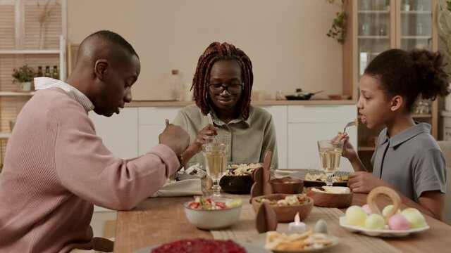 Steadicam young happy Black woman, her daughter and husband sitting at table on her sides, eating tasty festive dinner. Family celebrating Easter