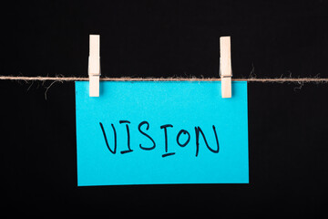 Vision word written on a Blue color sticky note hanging with a wire on black background.