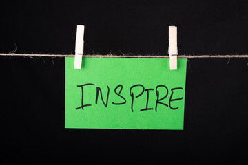 Inspire word written on a green color sticky note hanging with a wire on black background.
