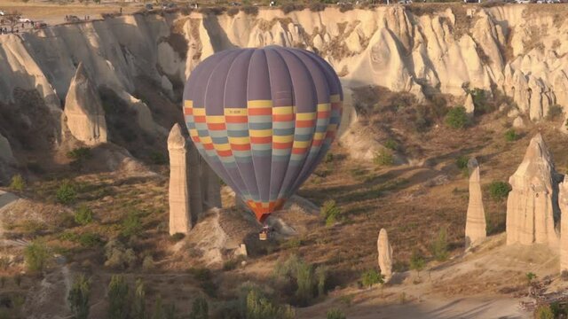 Colourful hot air balloon with brown tourist basket flies over large rocky cliffs of column shapes among rare green bushes at sunrise.