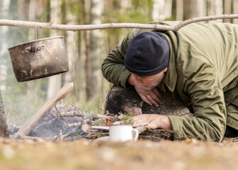 man in the forest trying to light a campfire, a tree branch with a pot over the campfire, blurred forest background, bonfire and smoke, autumn time