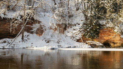 sunny day, landscape with red sandstone cliffs that are snowy with snow, frozen icicles on the cliff wall, icefall on the cliff wall, frozen river, Gauja, Kuku cliffs, Latvia
