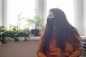 Red-haired curly girl in an orange jacket and black mask on her face sits near the window