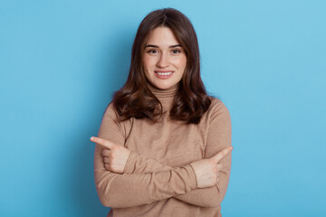 Obraz premium Portrait of happy cute girl pointing with fingers to different sides and looking at camera with cute shy smile, wearing beige turtleneck, posing isolated over blue background.