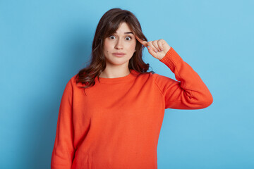 Portrait of dark haired woman in urban style jumper makes stupid sign with finger near head,...