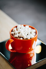 A cup of orange color with cocoa and marshmallows stands on a wooden table