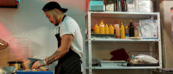 Side view of professional experienced sushi chef cutting ingredients with a knife for spreading them over nori sheet while making delicious rolls at commercial kitchen