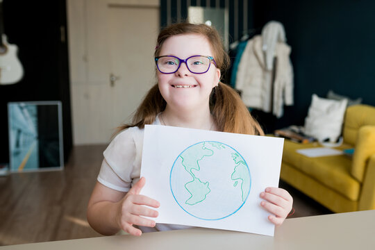 Portrait of teenage smiling girl with Down syndrome holding drawing of the planet Earth at home. Disabled child showing creative artwork. Down syndrome day.