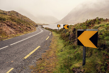 Small narrow asphalt road in a mountains by a lake, low clouds cover mountains peaks, fog...