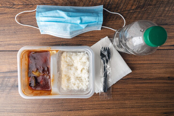 Fast food lunch on a wooden table with blue face mask, napkin, and plastic fork. Chinese style meal with white rice and bbq chicken in a plastic container