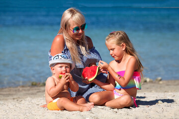 Mom and children are sitting on the beach near the sea and eating watermelon. Vacation.