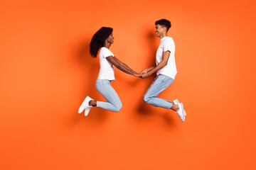Fototapeta na wymiar Full size profile side photo of young smiling afro couple on date holding hands jumping isolated on orange color background