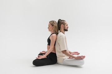 Fototapeta na wymiar А man and a woman sit and meditate on the floor with their backs against each other. Young people practicing contact yoga psychological therapeutic exercise in studio on white background.
