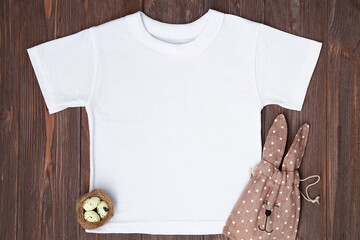 White t-shirt mockup with Easter eggs, nest and bunny candy bag flat lay on brown wooden background