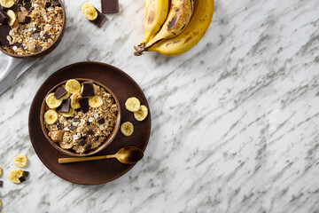  Natural muesli  in coconut bowl with  banana and chocolate on the marble table. Mix of unprocessed whole grains, chia, quinoa, nuts, seeds, fruit for heathy breakfast.