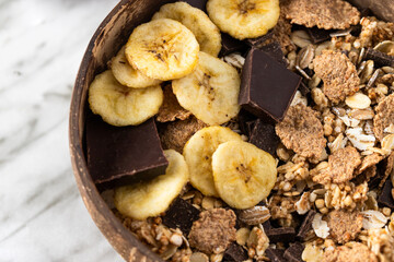  Close up of muesli  in coconut bowl with  banana and chocolate on the marble table. Mix of unprocessed whole grains, chia, quinoa, nuts, seeds, fruit for heathy breakfast.