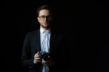 Portrait of a man wearing in black elegant suit looking at camera and holding photo camera, isolated on black background. Copy space.