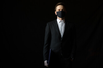 Businessman in medical mask and suit holding laptop while standing over black background with copy space. Concept of work, covid-19 and social distancing.