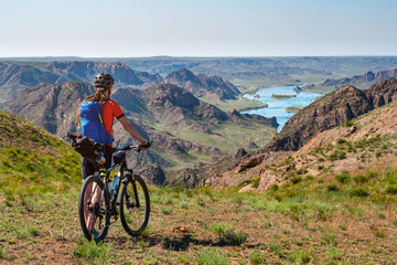 Fototapeta premium A traveler guy on a bicycle looks out over a beautiful river valley. A beautiful natural landscape of a river valley with hills and flowering meadows