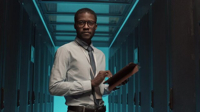 Medium shot portrait of young African-American man wearing formalwear standing at rack server cabinet, working on digital tablet and then looking at camera