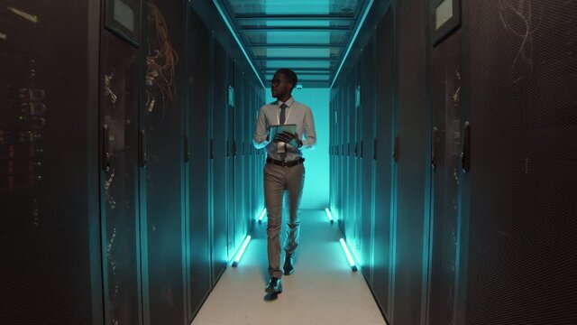 Lockdown of young African-American man wearing formalwear going along row of rack server cabinets and using digital tablet for maintenance of them