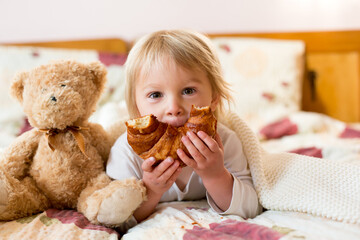Cute blonde toddler child,reading book and eating croissant in bed, laughing