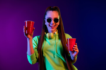 Photo of funky happy young girl hold red cups beer party clubber wear headset dj isolated on neon...