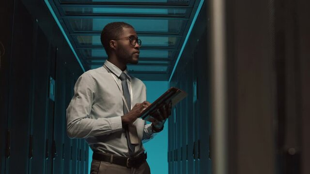 Medium shot of young African-American male IT technician standing in room with row of rack server cabinets and using digital tablet to maintain their work
