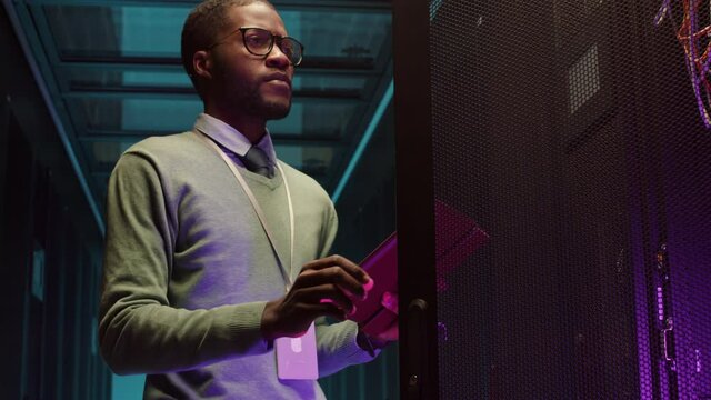 Low angle view of young African male IT administrator going along room with row of rack server cabinets, opening one of them and using tablet to analyze its work