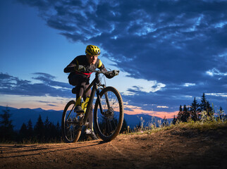 Fototapeta premium Back view of young man cycling bicycle under beautiful night sky. Male bicyclist in safety helmet riding on hillside road under blue cloudy sky in the evening. Concept of sport and active leisure.