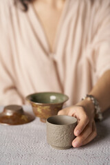 Cropped shot of lady with beige ceramic bowl with grained stone-like surface. The woman is sitting at the table with crockery ware and holding cylindric tea bowl in her hand. 
