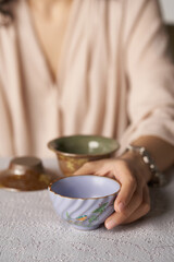 Fototapeta na wymiar Cropped shot of lady with violet ceramic bowl with glazed surface, crazing and floral Japanese design. Woman is sitting at the table with crockery ware and holding small wavy tea bowl in her hand. 