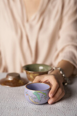 Fototapeta na wymiar Cropped shot of lady with violet ceramic bowl with glazed surface, crazing and floral Japanese design. Woman is sitting at the table with crockery ware and holding small tubby tea bowl in her hand. 