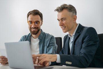 Fototapeta premium Successful software engineers using laptop, talking, cooperation working together in office. Smiling business people talking, searching online, meeting, planning start up sitting at workplace 