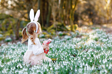 Adorable little girl with Easter bunny ears eating chocholate figure in spring forest on sunny day, outdoors. Cute happy child with lots of snowdrop flowers. Springtime, christian holiday concept.