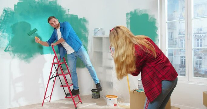 Rear of Caucasian woman standing in room taking photos on smartphone of her young handsome husband posing and painting wall in green color in good mood. Home repair, renovation and makeover concept
