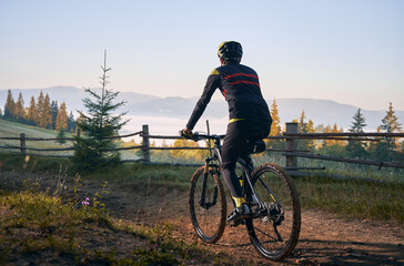 Back view of male cyclist riding bike on mountain road with coniferous trees and hills on background. Man bicyclist enjoying bicycle ride in mountains. Concept of sport, biking and active leisure.