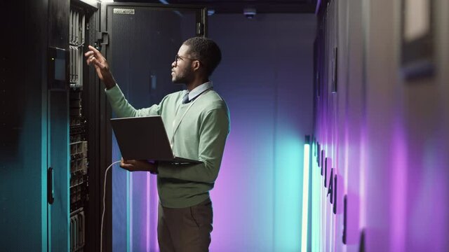Lockdown of young African-American male IT technician checking work of hardware in data center illuminated with neon light using laptop