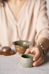 Cropped shot of lady with beige ceramic bowl with grained surface and glazed inner side. The woman is sitting at the table with crockery ware and holding tubby tea bowl in her hand. 