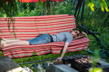 A man with glasses in the summer sleeps on a swing, bench, campfire and fries meat