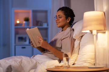 people, bedtime and rest concept - happy smiling young african american woman in glasses reading book in bed at home at night