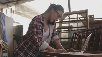 Asian woman carpenter wearing safety eyes goggle and talking to customer by smartphone in workplace. Young female carpentry using computer laptop and mobile phone for customer order in small factory.  - Powered by Adobe