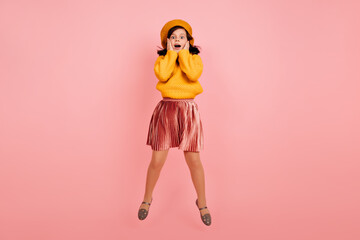 Full length shot of amazed child in yellow beret. Little brunette girl jumping on pink background.
