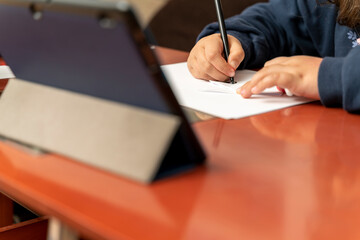 detail of a girl's hands drawing or writing on a sheet of paper while she listens to the explanations through a tablet. Concept of distance education and social distance.