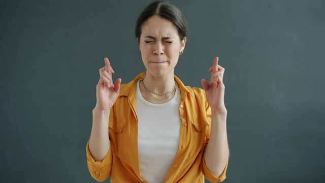Portrait of beautiful girl holding fingers crossed and praying gesture standing on gray background. Hopeful young people and good luch concept.