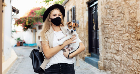 tourist in protective mask holds dog on hands and walking in old city Coronavirus disease COVID-19 new travel reality. Girl in medical mask walking. Coronavirus pandemic Precautions. Travel with pets