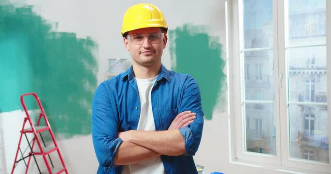 Portrait of Caucasian handsome happy young man repairman in yellow casque stand in room during home renovation process looking at camera and smiling, job profession reconstruction construction concept