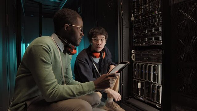 Lockdown of male African IT specialist and his Asian coworker squatting at rack server cabinet in data center and controlling its work together
